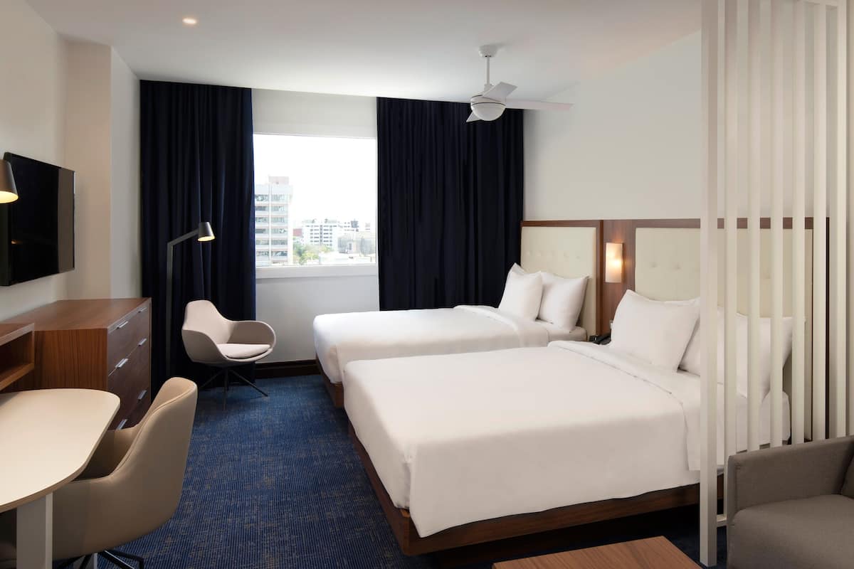 Ein Hotelzimmer mit zwei Betten, einem Schreibtisch, einem Stuhl, einem Fernseher und einem Fenster mit Vorhängen.