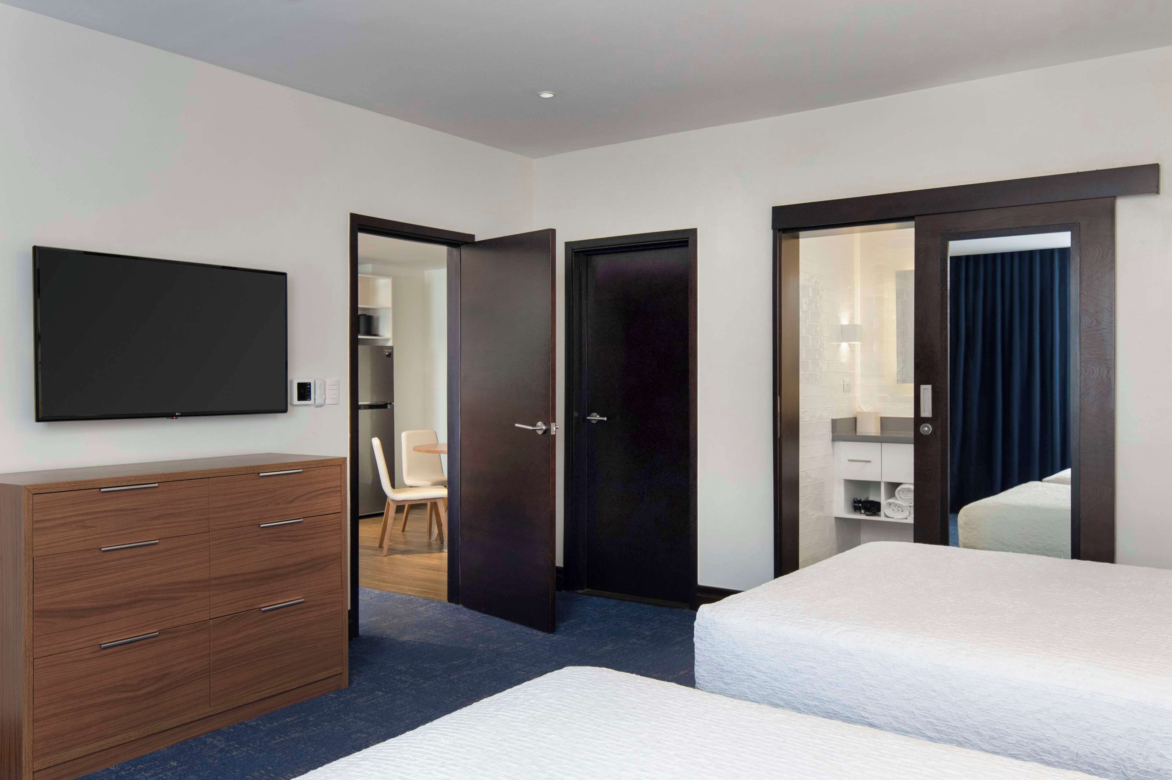 Quarto de hotel com uma cama, cômoda, televisão e banheiro visível por uma porta aberta.