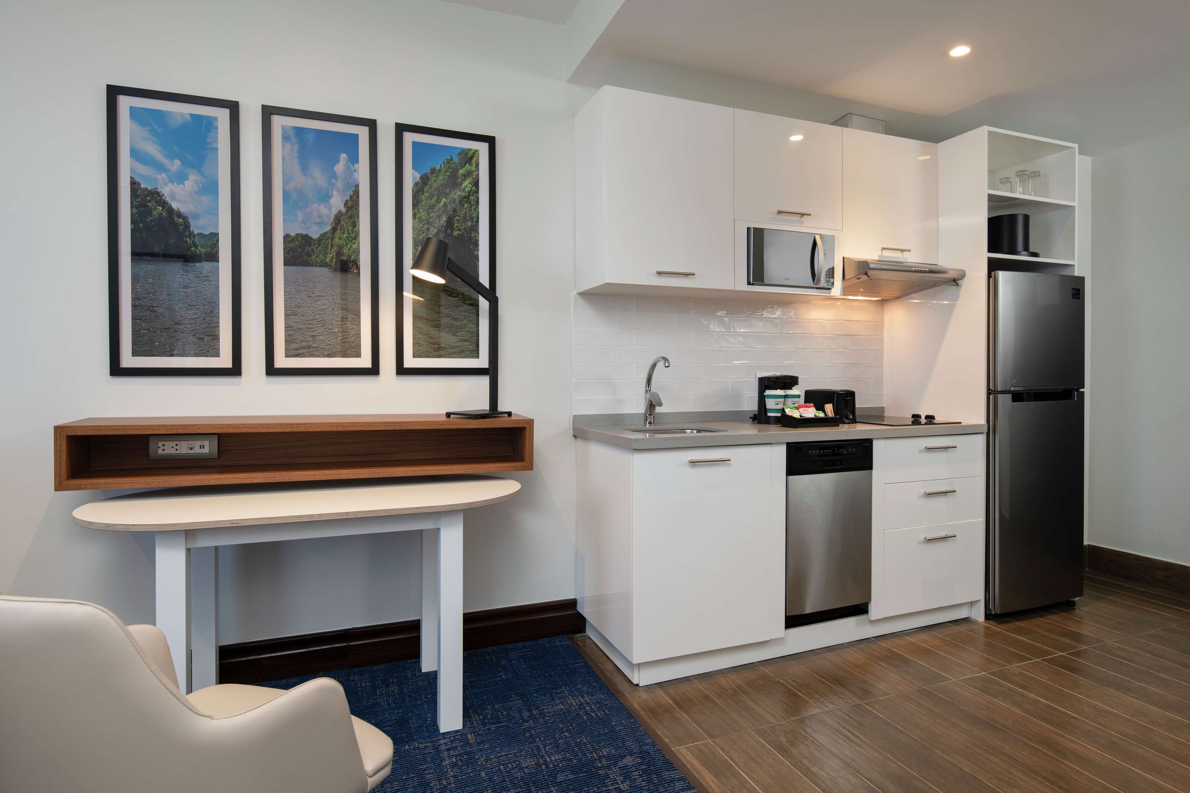 Cocina moderna con armarios blancos, un refrigerador de acero inoxidable, un microondas y un lavavajillas. Hay una mesa pequeña con una silla, y tres cuadros en la pared.