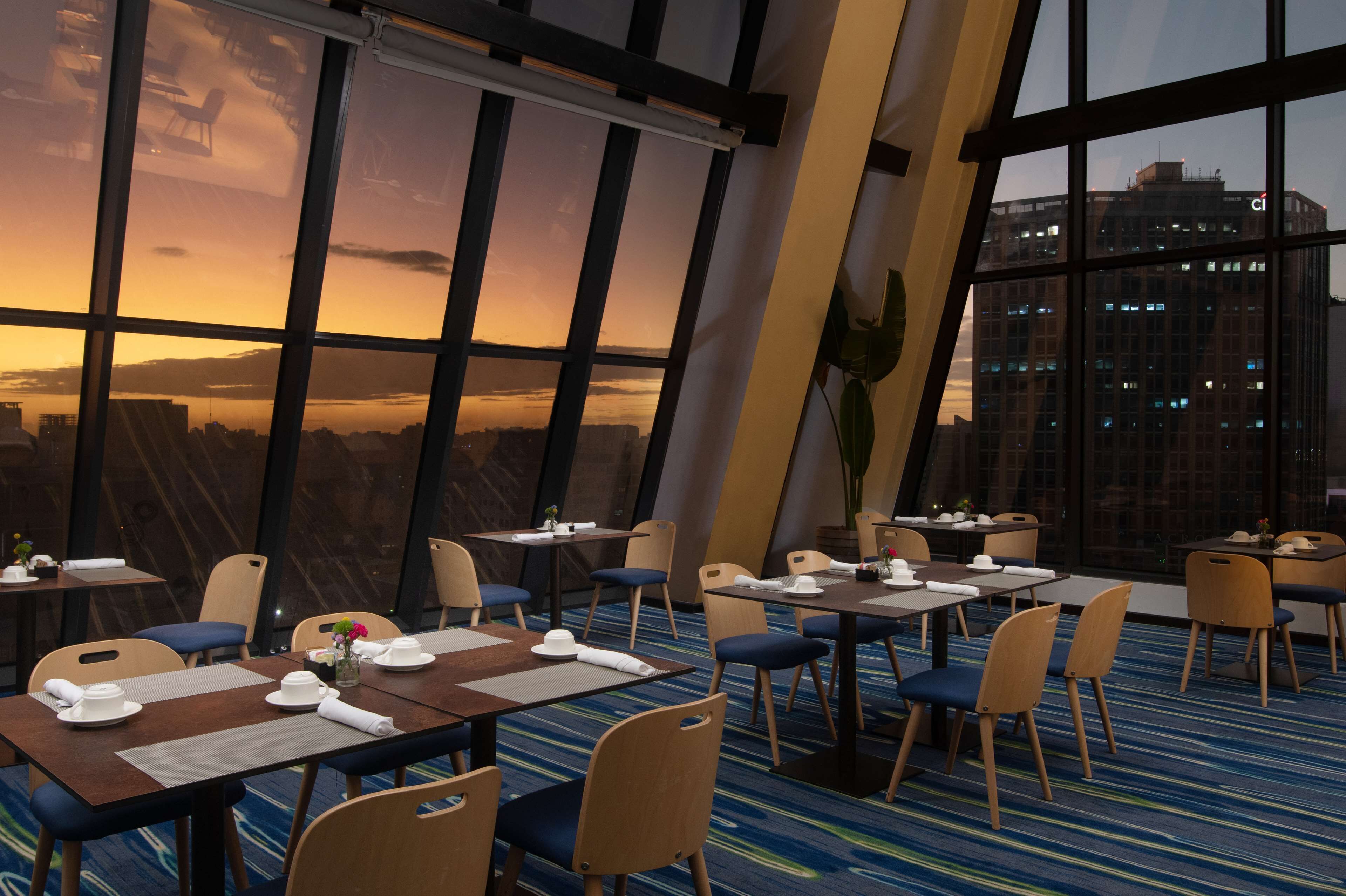 A restaurant with tables and chairs arranged for dining, large windows offering a view of the cityscape, and a modern interior design.