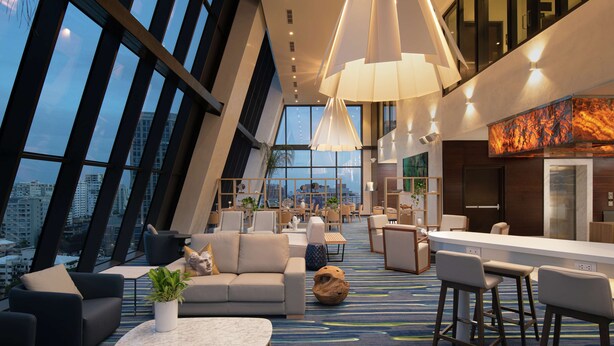 Un vestíbulo de hotel moderno con techo alto, ventanas amplias y una zona de bar central.