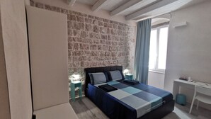 Un dormitorio con pared de piedra, una cama con colcha a cuadros, una mesita de noche con lámpara y un escritorio con silla.