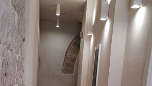 Un pasillo con una pared de piedra a un lado, paredes color beige y piso de baldosas. Hay lámparas en las paredes y una escalera al fondo.