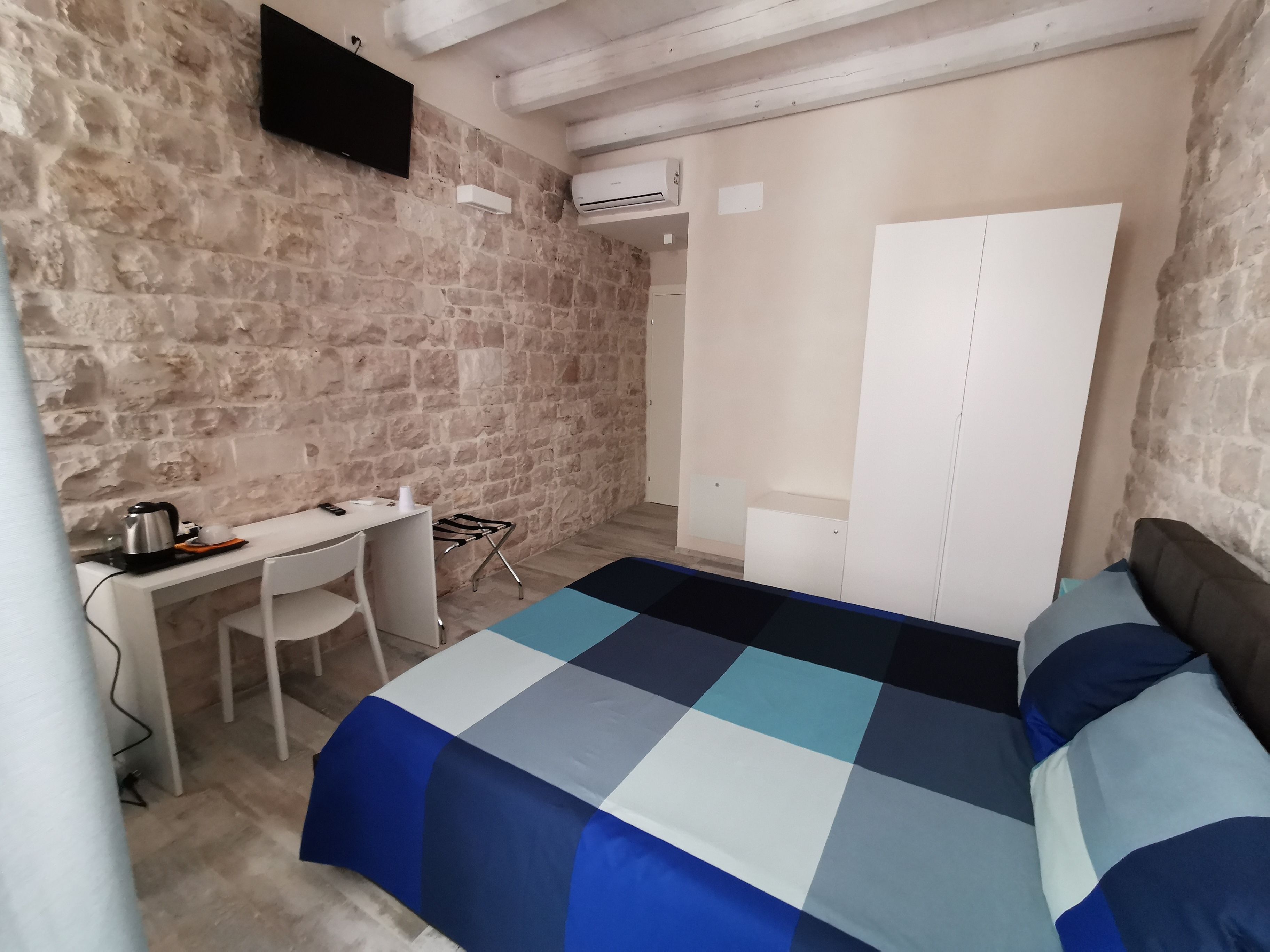 A bedroom with a stone wall, a bed with a blue and white checkered bedspread, a desk with a kettle, and a chair.