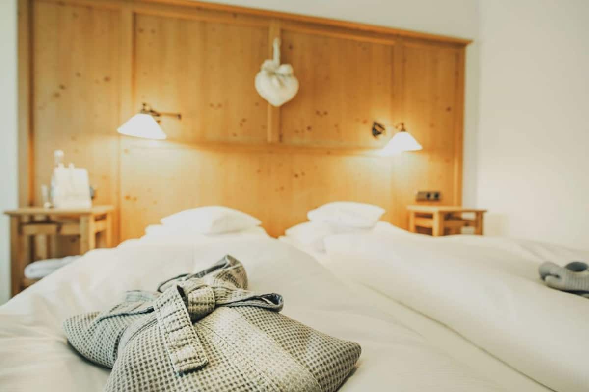A hotel room with a wooden headboard, two bedside lamps, a bed with white linens, and a small bedside table with a bottle and a glass.