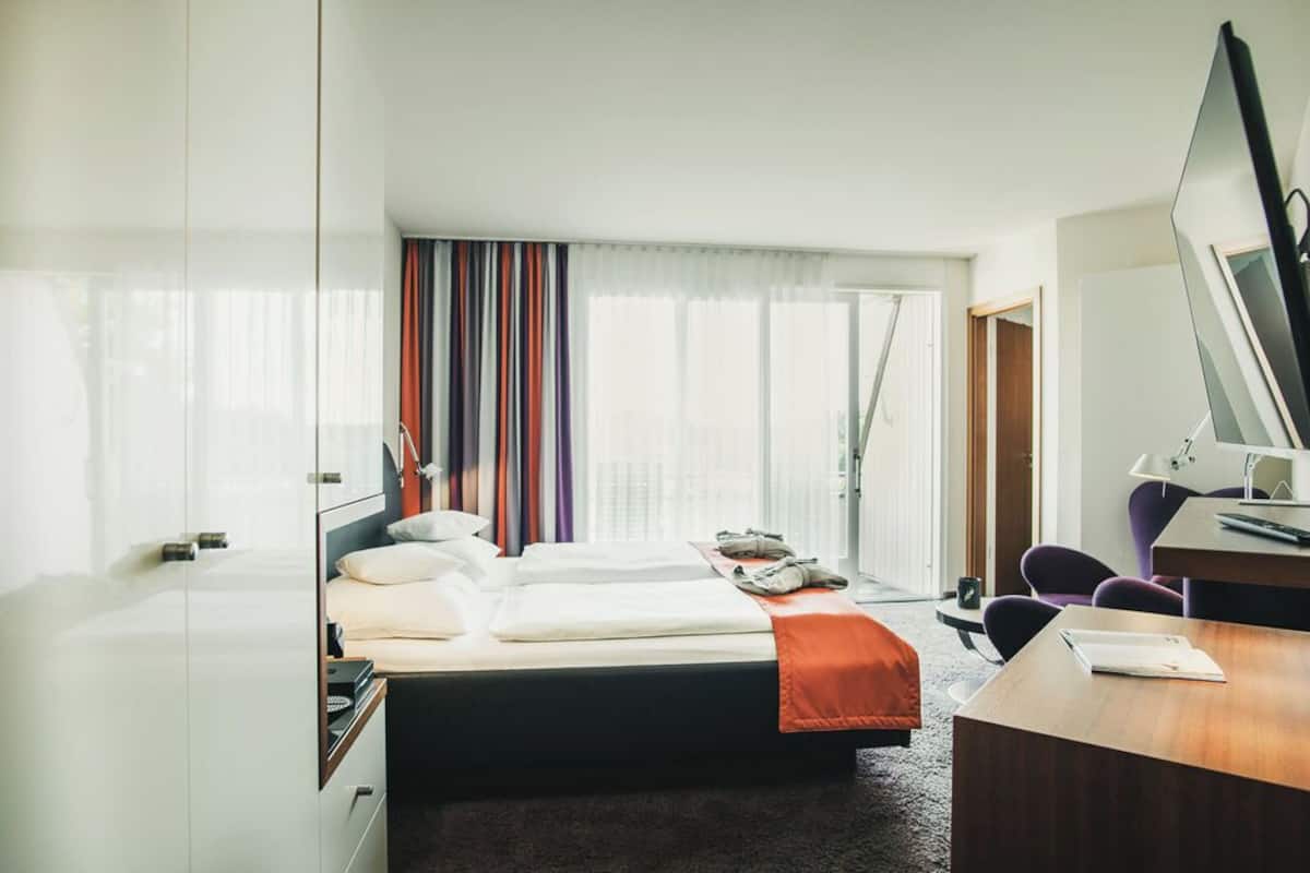 A hotel room with a bed, a desk, a chair, a wardrobe, and a large window with curtains.