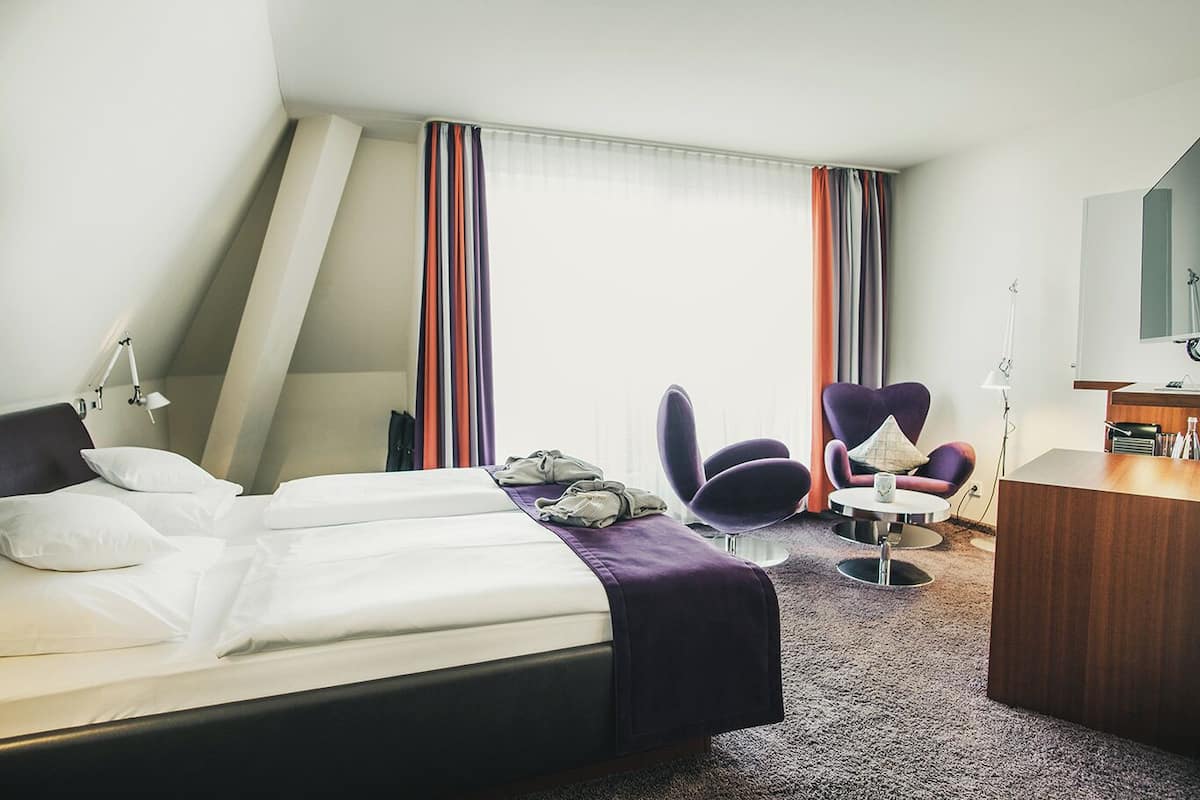 A hotel room with a bed, two armchairs, a desk, and a large window with curtains.