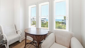A room with a white chair, a round table, and a sofa. There are three windows with blinds, and a view of a building and greenery outside. - Welcome to Sea La Vie! (St. George Island)
