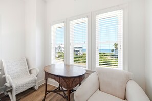 A room with a white chair, a round table, and a sofa. There are three windows with blinds, and a view of a building and greenery outside.