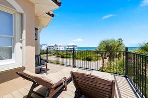 A balcony with two lounge chairs, a view of the ocean, and a paved walkway with a fence.