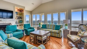 Un salon spacieux avec des canapés turquoise et à rayures, une table basse en bois et une cheminée. De grandes fenêtres offrent une vue sur l’extérieur.