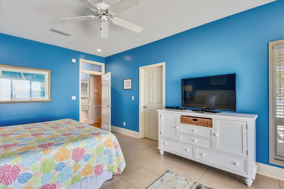 A bedroom with a bed, a television on a white dresser, a ceiling fan, and a door leading to another room.