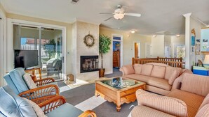 A living room with a fireplace, a television, wicker furniture, and a ceiling fan. - TRADEWINDS HAS LONG BEEN praised as a uniquely appointed home where treasured memories are made. (St. George Island)