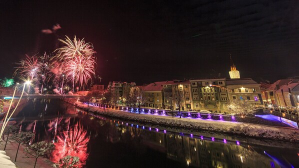 Vista nocturna de una zona ribereña con fuegos artificiales, edificios iluminados y un puente.