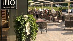 An outdoor seating area with tables and chairs, a sign for 'MiLo Alpen Adria Cuisine', and a view of mountains in the background. - Hotel Seven (Villach)