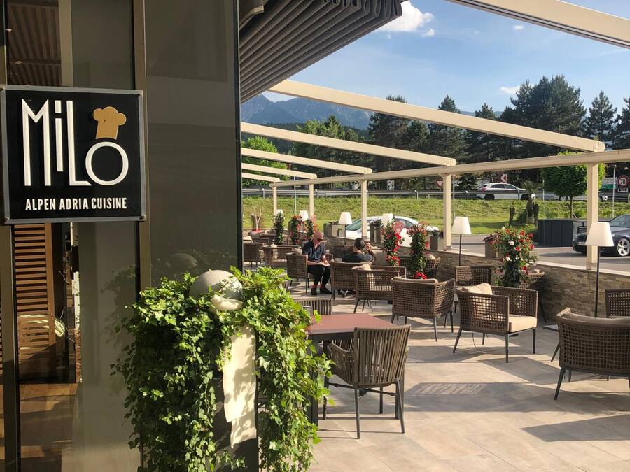 Un área de estar al aire libre con mesas y sillas, un cartel que dice 'MiLo Alpen Adria Cuisine' y una vista de montañas al fondo.