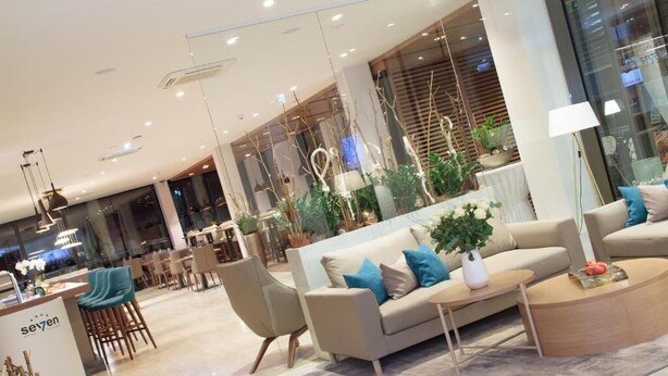 A modern hotel lobby with a seating area featuring a sofa and chairs, a reception desk, and a dining area in the background. - Hotel Seven (Villach)