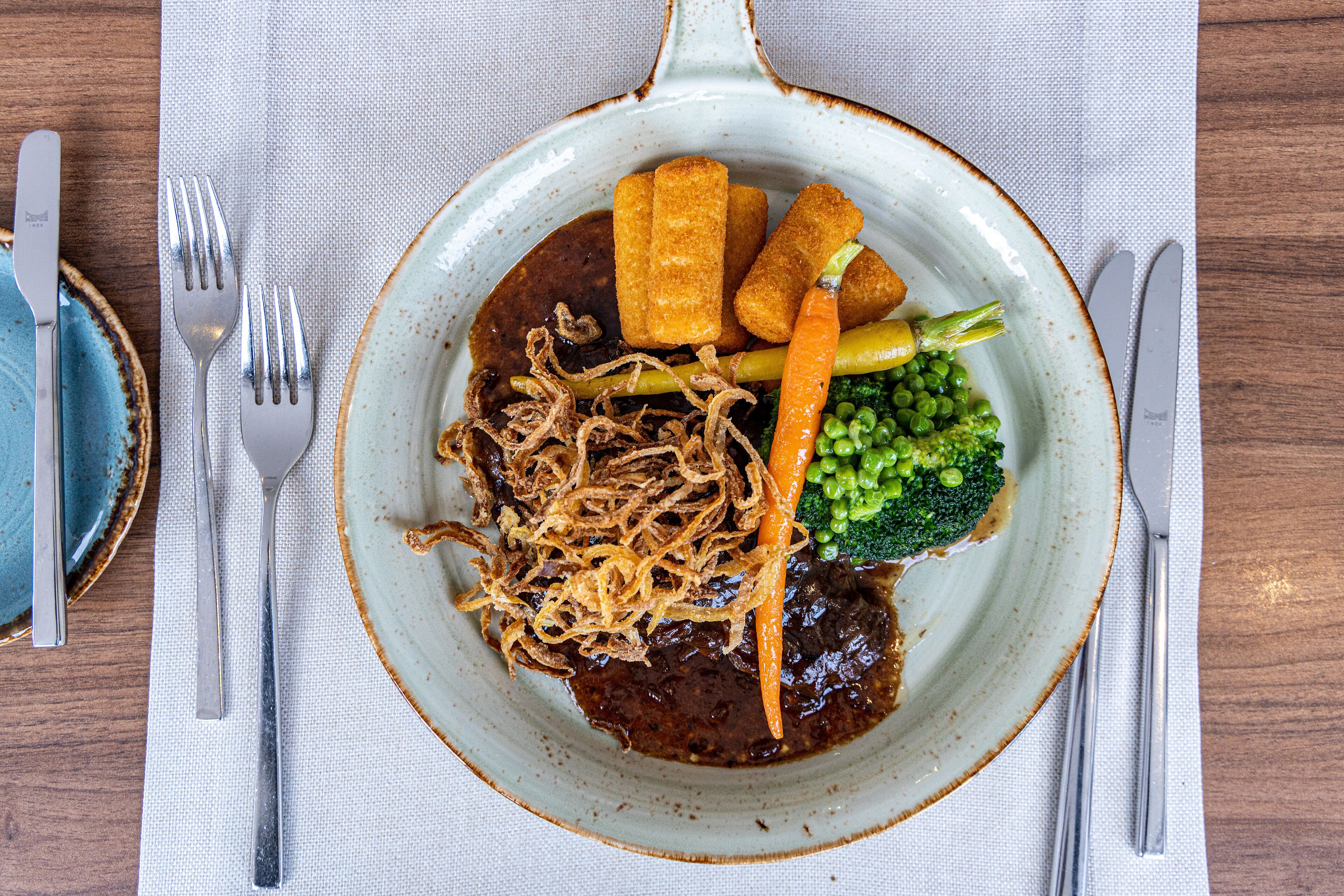 A plate of food with various vegetables and a meat dish, accompanied by cutlery on a tablecloth.