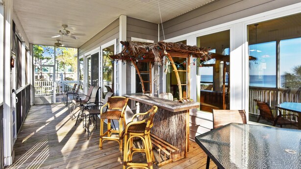 A covered outdoor area with a wooden deck, wicker chairs, a bar area, and large windows offering a view of the ocean.