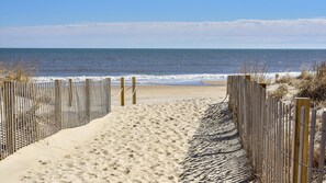Un sentier de sable qui longe la plage et mène à l’océan, bordé de clôtures en bois et de quelques plantes.