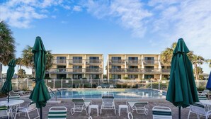 A poolside area with lounge chairs and umbrellas, a pool, and a multi-story building in the background. - Located right on the beachfront, this beautiful well-appointed Villa offers you fabulous views. (St. George Island)