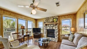 A spacious living room with a stone fireplace, wooden walls, and large windows offering a view of the outdoors. - DELIGHT IN THE CHEERFUL AMUSEMENT of the sand crabs dancing in the moonlight. (St. George Island)