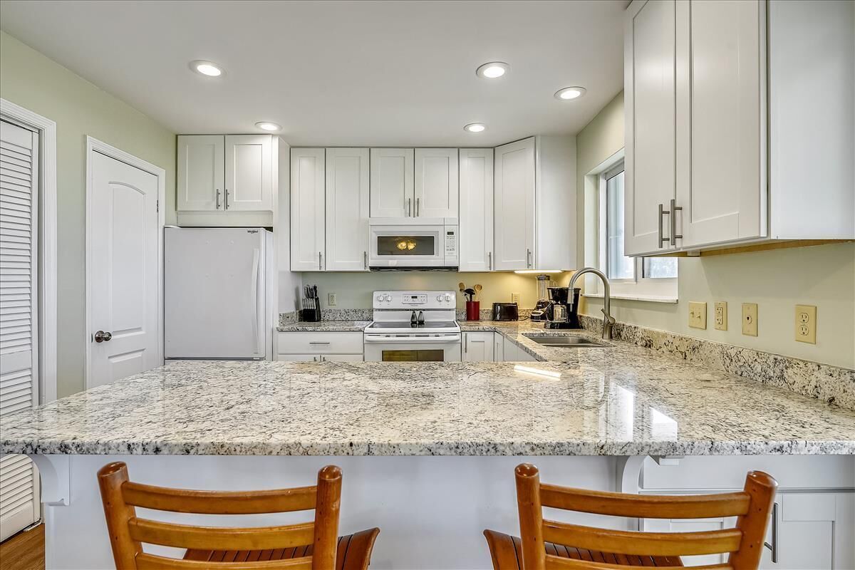Cocina con gabinetes blancos, encimera de granito, microondas, estufa y dos sillas de madera.