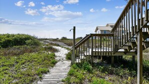 A wooden boardwalk leading to a wooden staircase with a railing, surrounded by greenery and a clear sky. - DELIGHT IN THE CHEERFUL AMUSEMENT of the sand crabs dancing in the moonlight. (St. George Island)