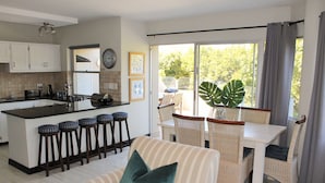 A modern kitchen with a central island, bar stools, a dining area with wicker chairs, and a large window with curtains. - Lagoon Terrace 14 (Plettenberg Bay)