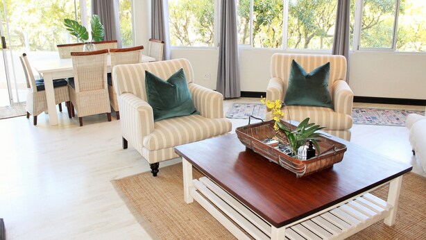 A spacious living room with a dining area, large windows with curtains, and a wooden coffee table with decorative items. - Lagoon Terrace 14 (Plettenberg Bay)