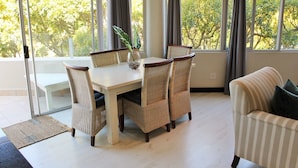A dining area with a table and chairs, a vase with a plant, and large windows with curtains. - Lagoon Terrace 14 (Plettenberg Bay)
