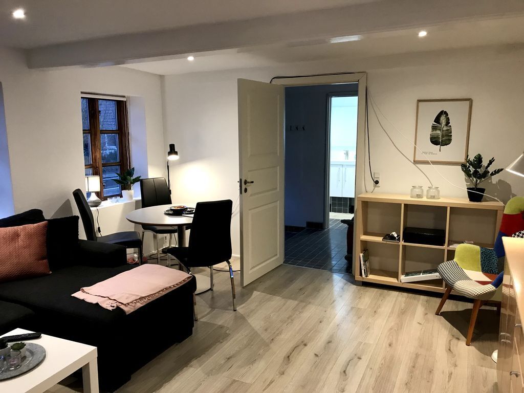 A modern living room with a black sofa, a dining table with chairs, a wooden bookshelf, and a door leading to another room.