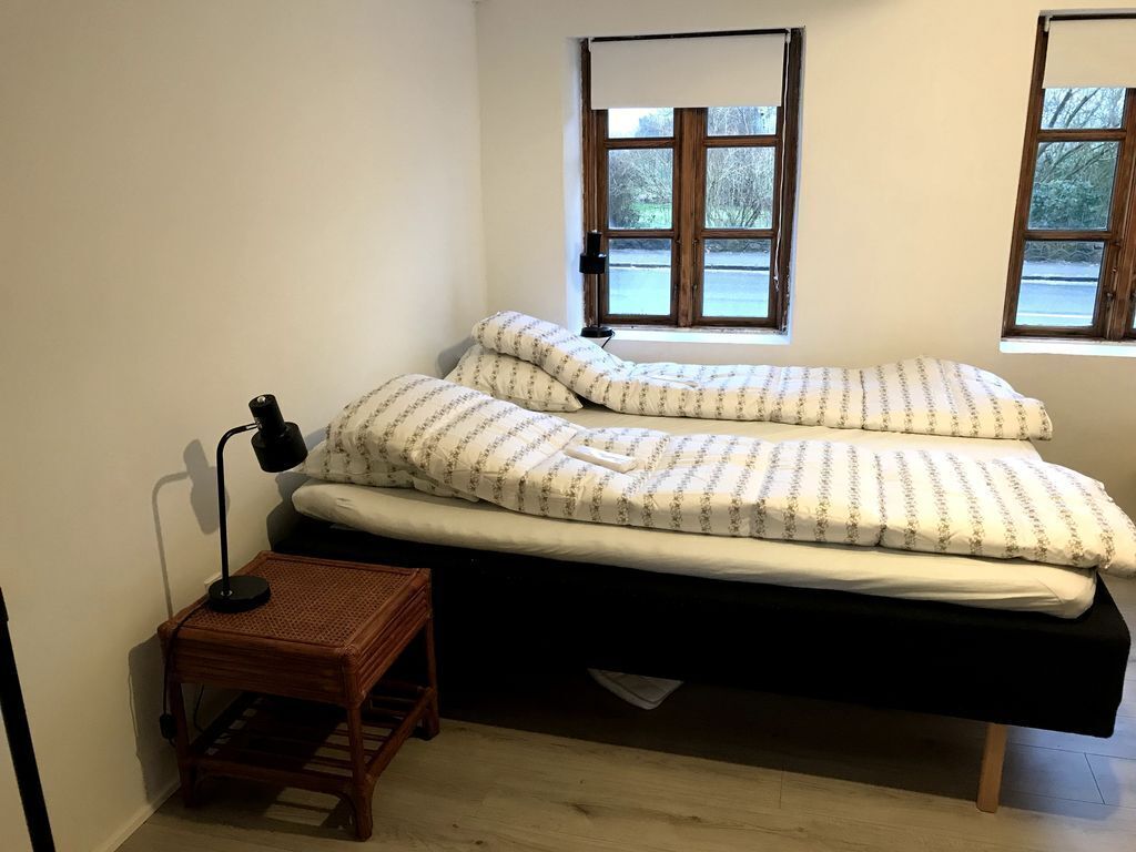 A small, single-bed room with a wooden floor, a bedside table with a lamp, and two windows with wooden frames.