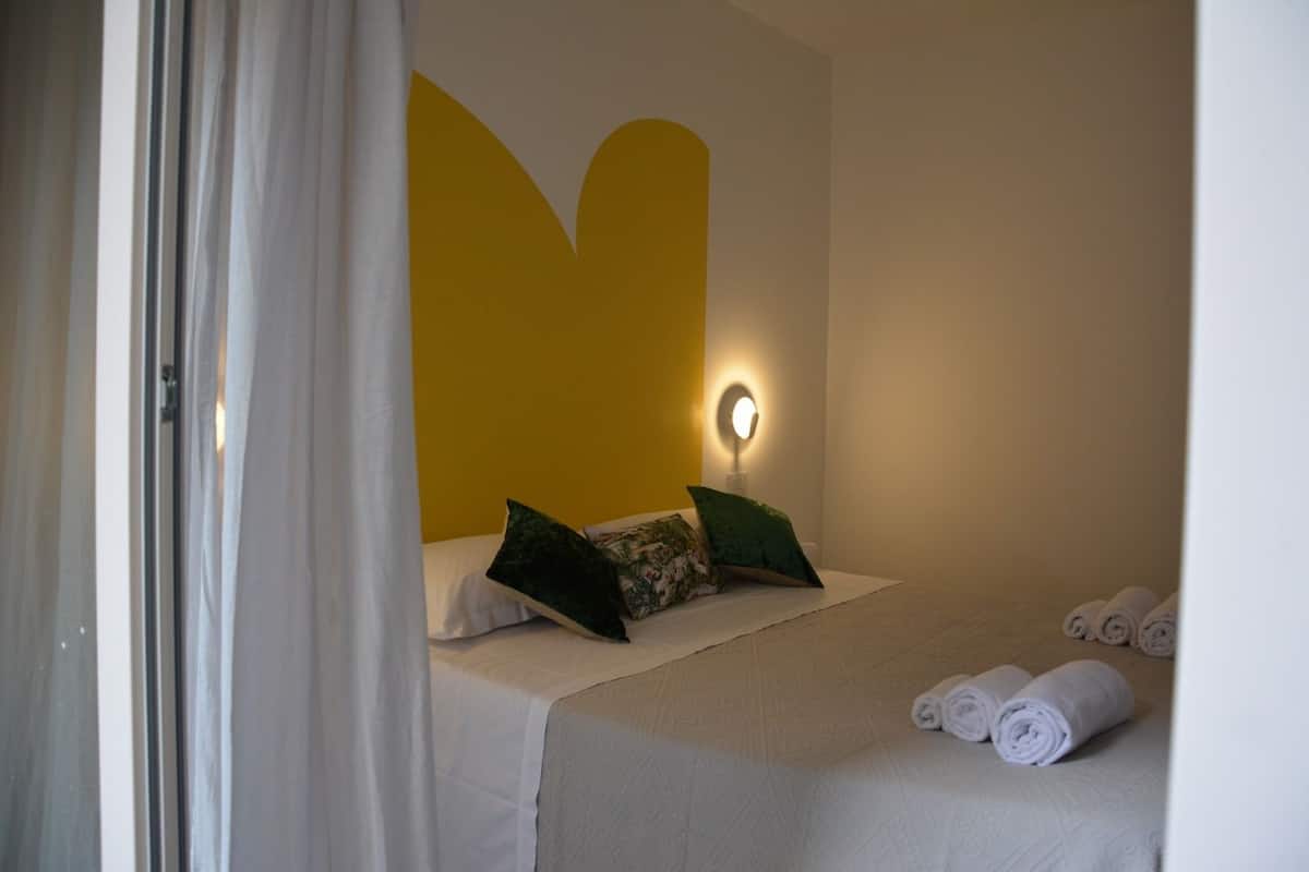 Une chambre d’hôtel avec un lit, des oreillers, une lampe fixée au mur et des serviettes enroulées posées sur le sol.