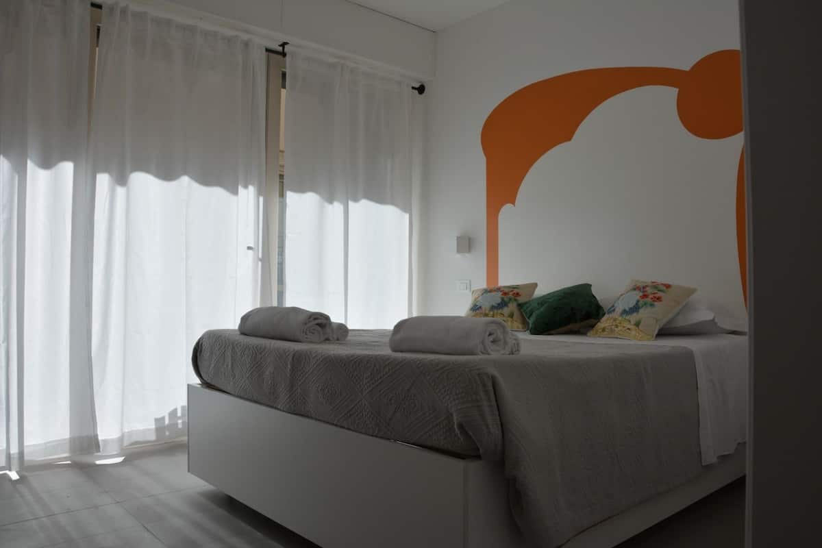 Une chambre moderne avec un grand lit, des rideaux blancs et un mur orné d’un grand motif abstrait orange.