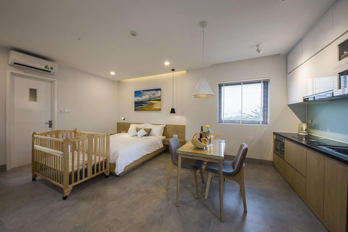 A modern hotel room with a crib, a bed, a dining table with chairs, a kitchen area, and a painting on the wall.