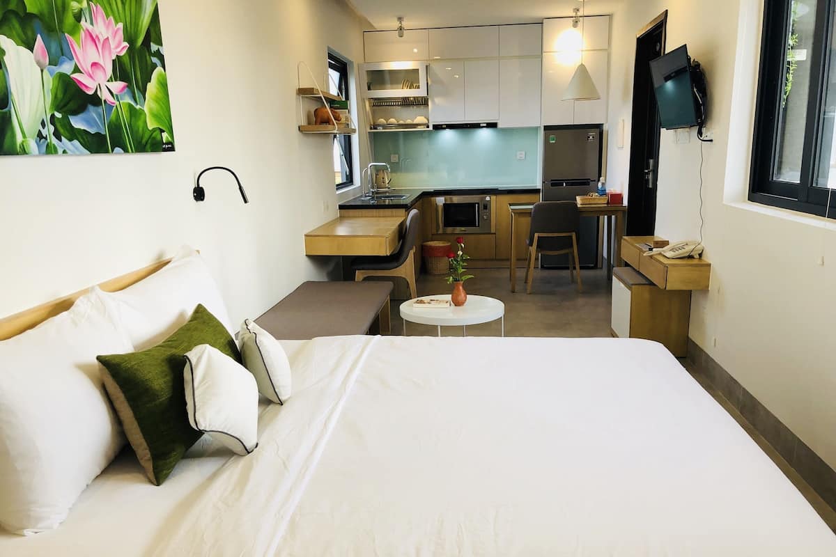 A hotel room with a bed, a small table with a vase, a desk, a chair, a kitchen area, and a TV mounted on the wall.