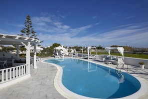 Un área de piscina con una piscina curva, sillas reclinables blancas y una pérgola.