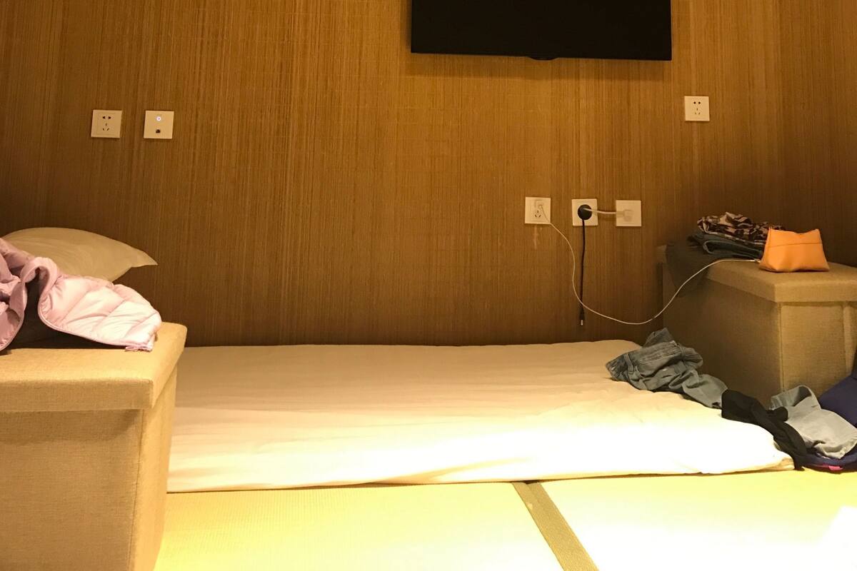 A hotel room with a wooden panel wall, a flat-screen TV mounted above a bed, and a bedside table with a lamp.