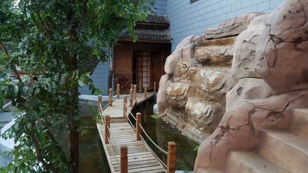 A wooden bridge over a water feature, a traditional building with a tiled roof, and a rocky wall. - Beijing Eastern Library Hotel (Huairou)