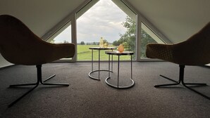 Una habitación con techo inclinado, dos sillas, una mesita con un florero y vistas a un campo verde a través de amplias ventanas.