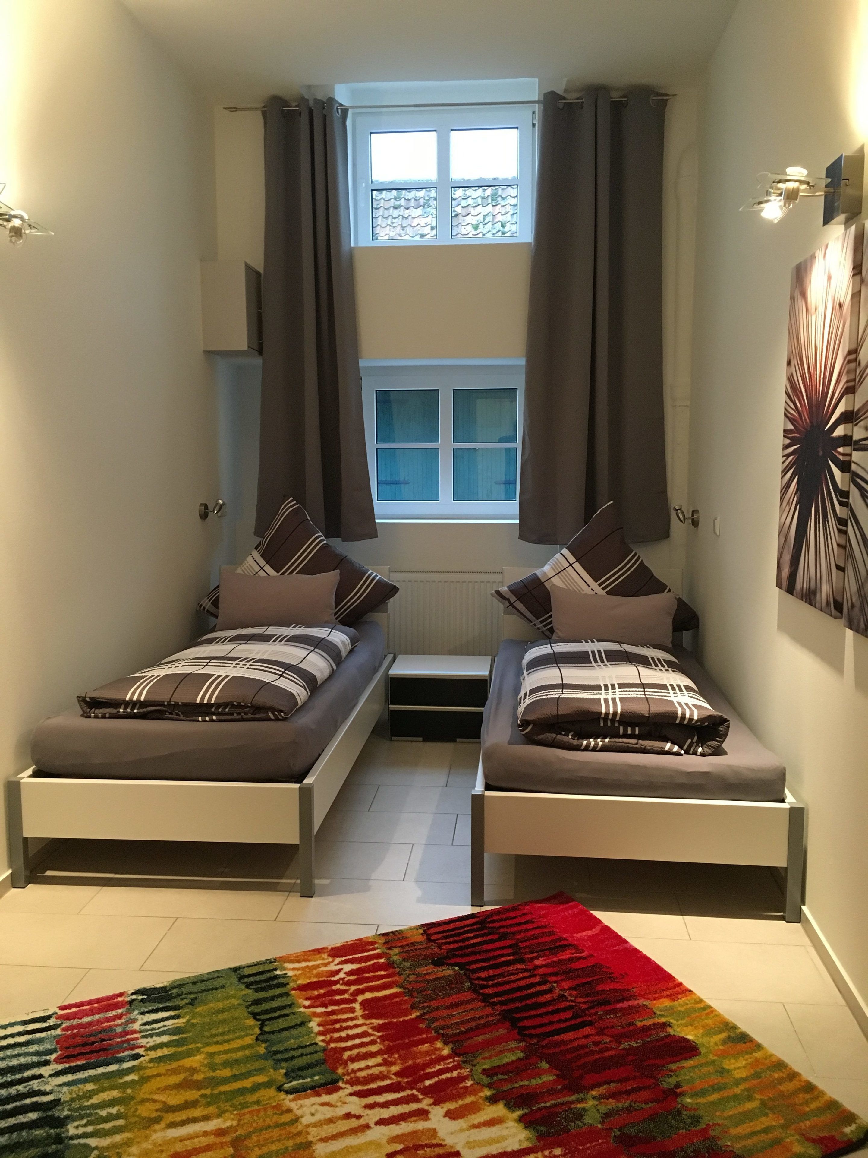 A small, well-lit room with two beds, a window with curtains, a radiator, and a colorful rug.