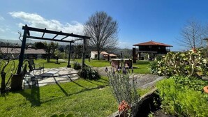 A patio with a pergola, a stone table, and a bench. There are trees and a building with a red roof in the background. - Hotel y Apartamentos Son de Mar (Villaviciosa)