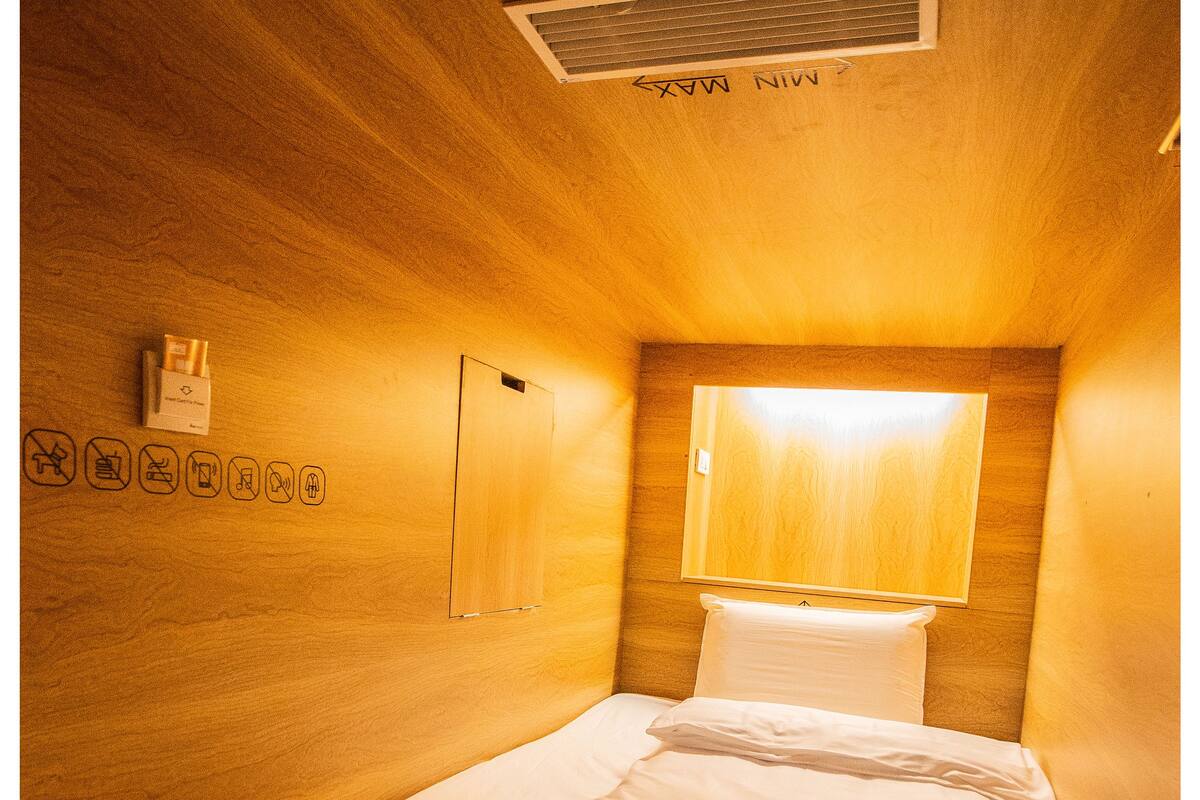 A small, cozy room with a single bed, a nightstand, a wall-mounted lamp, and a vent.