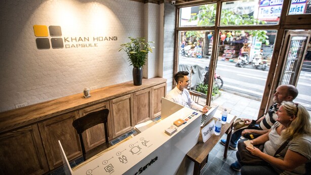 Un comptoir d’accueil orné du logo de « KHAN HOANG CAPSULE », un comptoir en bois, une plante en pot et une fenêtre donnant sur la rue.