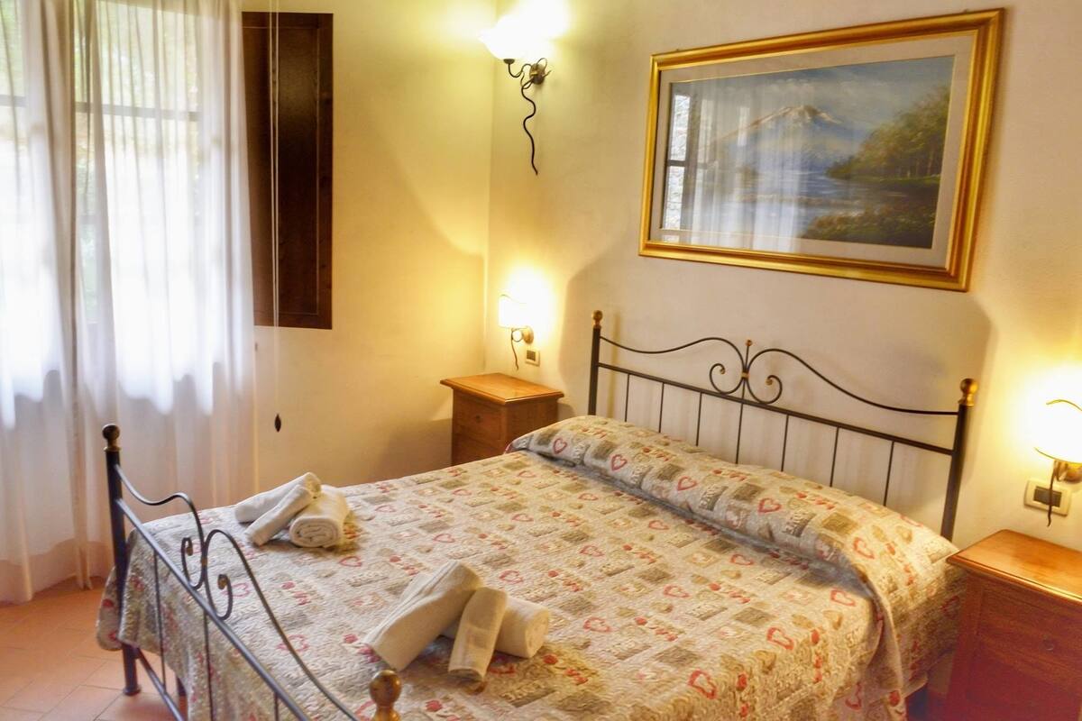 Una cama bien hecha con una colcha estampada y toallas enrolladas, una mesita de noche con una lámpara, un cuadro en la pared y una ventana con cortinas translúcidas.