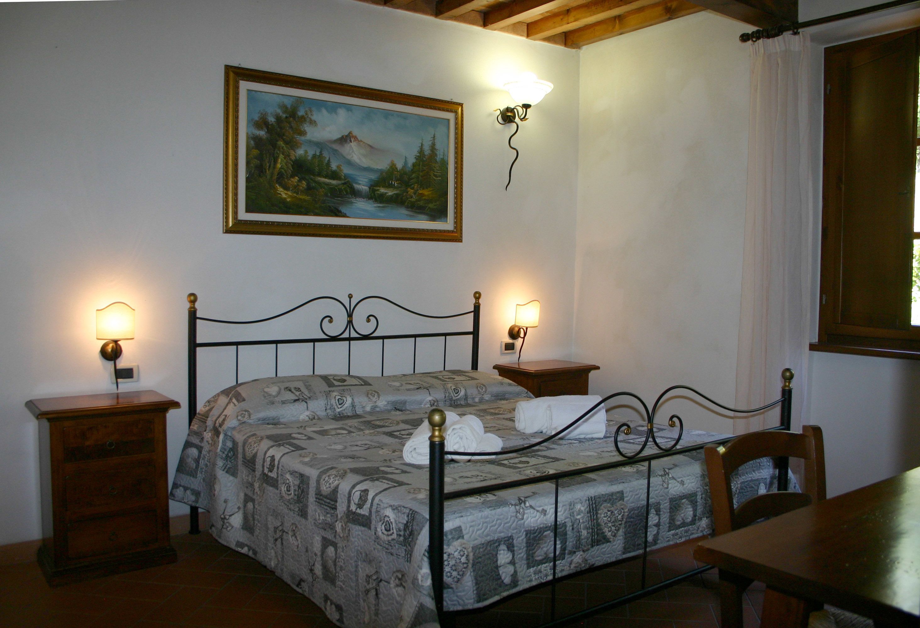 Quarto com cama, duas mesas de cabeceira, mesa de apoio, cadeira de madeira, mesa e um quadro na parede, além de uma janela com cortinas.