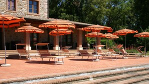 Zona junto a la piscina con varios sillones de sol y sombrillas naranjas, rodeada por un edificio de piedra con un sendero de ladrillo que lleva a la piscina.