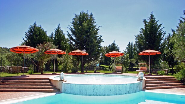Piscina con sombrillas rojas, rodeada de árboles y un solárium de ladrillo.