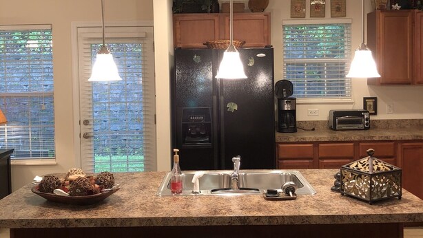 A kitchen with a central island, three bar stools, a refrigerator, a coffee maker, and pendant lights.